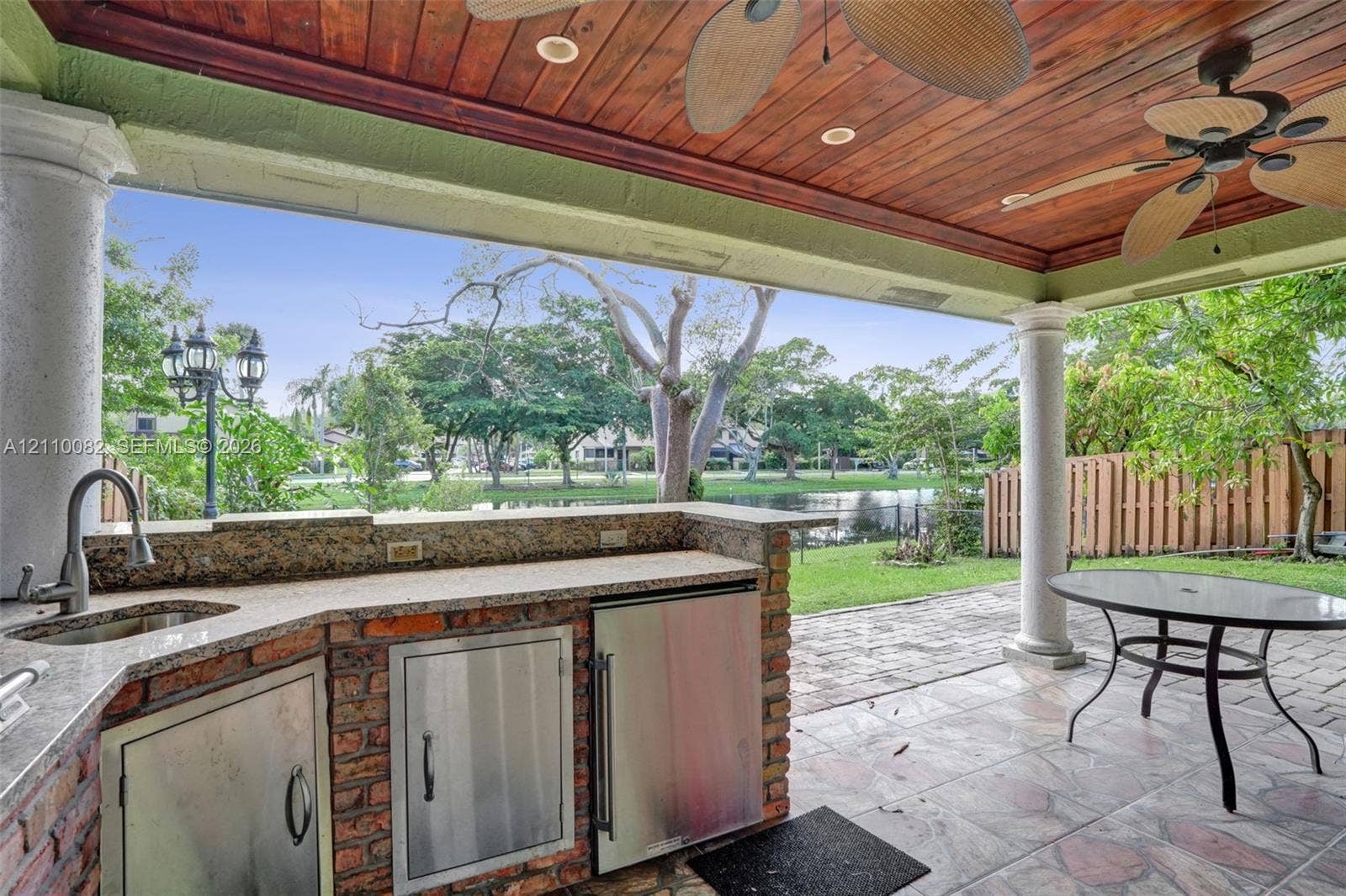 Covered outdoor kitchen featuring brick construction, stone countertop, stainless steel cabinetry, and undermount sink, complemented by a wood-plank ceiling, ceiling fans, and stone-tiled patio