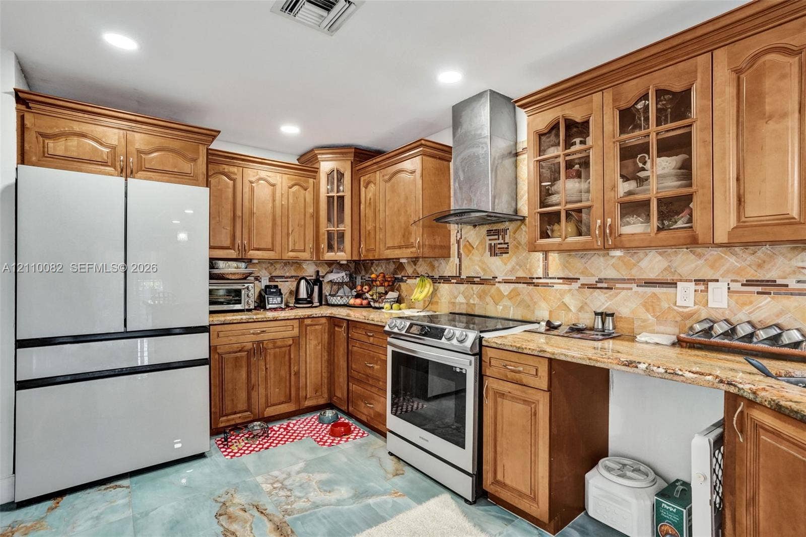 Kitchen featuring stained wood cabinetry with glass-front display accents, stone-finish countertops, herringbone-patterned tile backsplash, stainless steel range with a matching chimney-style hood, and large-format decorative floor tile