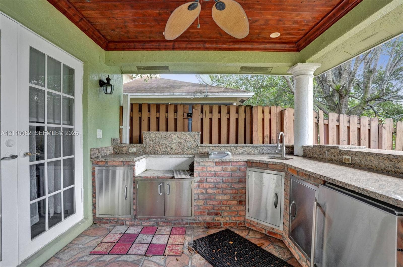 Covered outdoor kitchen featuring a brick-base structure, natural stone countertops, integrated stainless steel cabinetry, a built-in sink, and a stained wood-plank ceiling with a ceiling fan