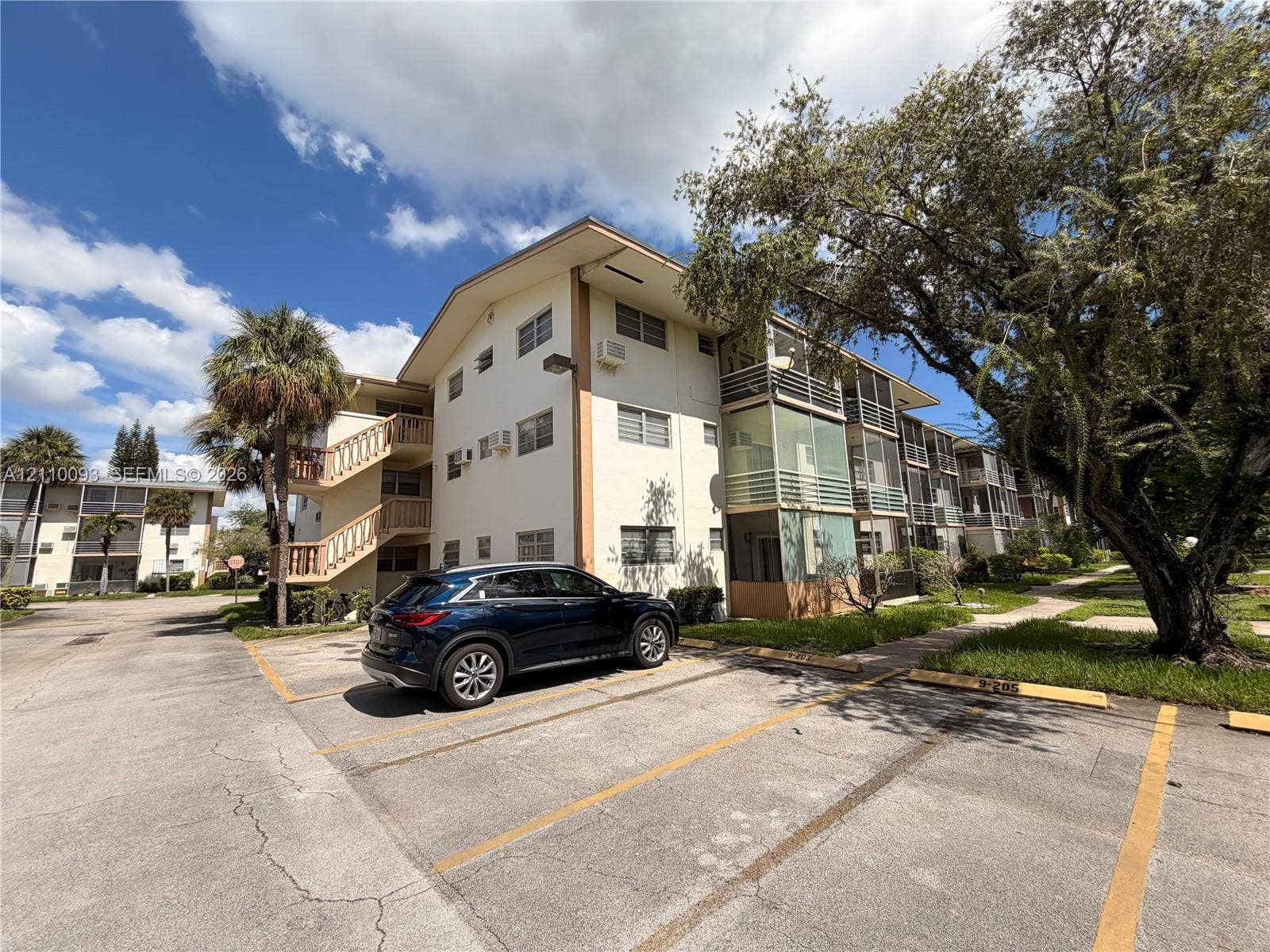 1395 NE 167th Street, #205, Miami, FL 33162 Multi-story exterior featuring a light-colored stucco facade, railed staircases, screened-in balconies, and designated asphalt parking