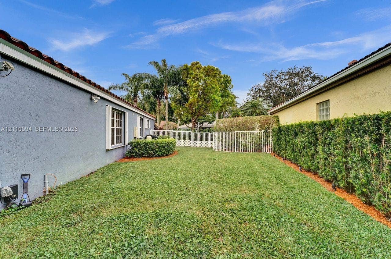 2064 Island Circle, Weston, FL 33326 Side yard featuring a grass lawn, metal fencing, stucco exterior walls, and manicured hedge landscaping