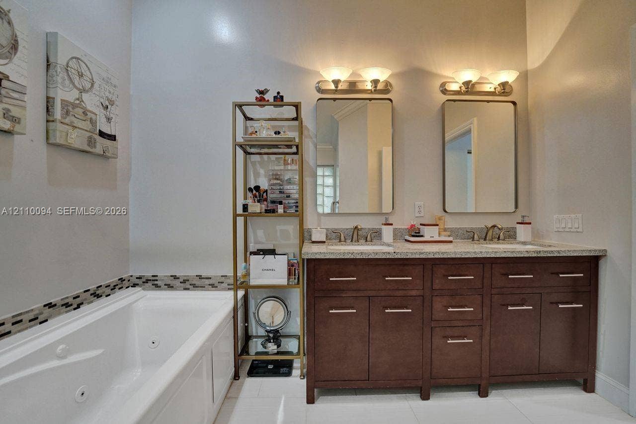 2064 Island Circle, Weston, FL 33326 Double-sink vanity featuring dark wood-finish cabinetry, stone-composite countertop, dual rectangular mirrors, and polished metal lighting fixtures, paired with a jetted soaking tub featuring an integrated tile backsplash