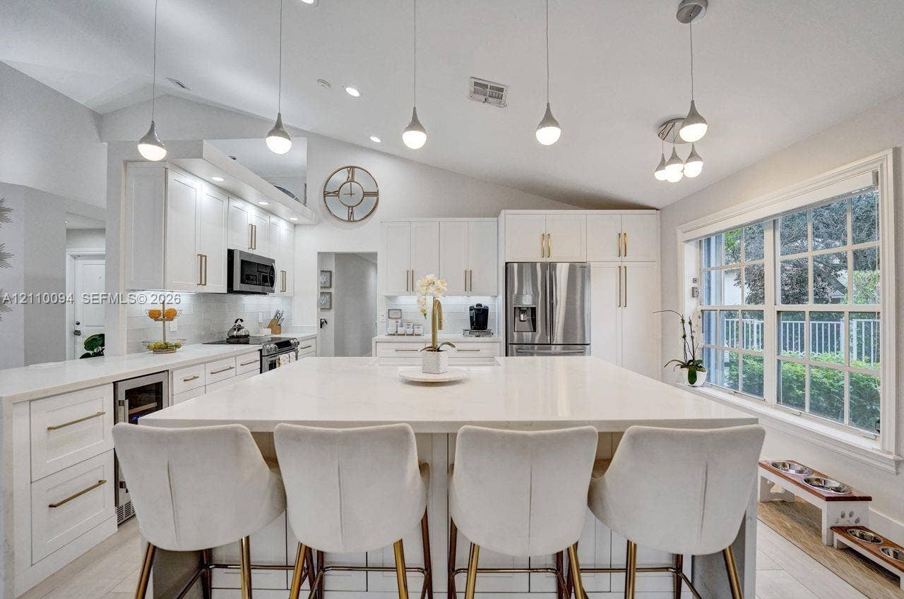 2064 Island Circle, Weston, FL 33326 Kitchen featuring white cabinetry with gold-tone hardware, a large center island with a stone-finish countertop, stainless steel appliances, and vaulted ceilings with multiple pendant light fixtures