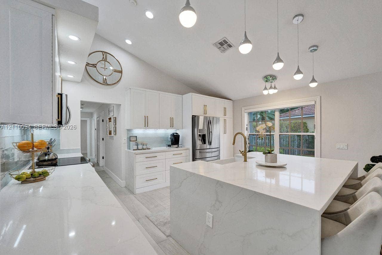 2064 Island Circle, Weston, FL 33326 Kitchen featuring white cabinetry with gold-tone hardware, waterfall edge island with stone-finish countertop, under-cabinet lighting, vaulted ceilings, and pendant light fixtures