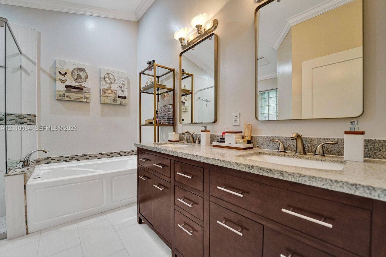 2064 Island Circle, Weston, FL 33326 Dual-sink vanity with dark wood-finish cabinetry, stone-composite countertops, rectangular undermount sinks, and metallic hardware, featuring a deep soaking tub with a tiled surround and ceramic floor tile