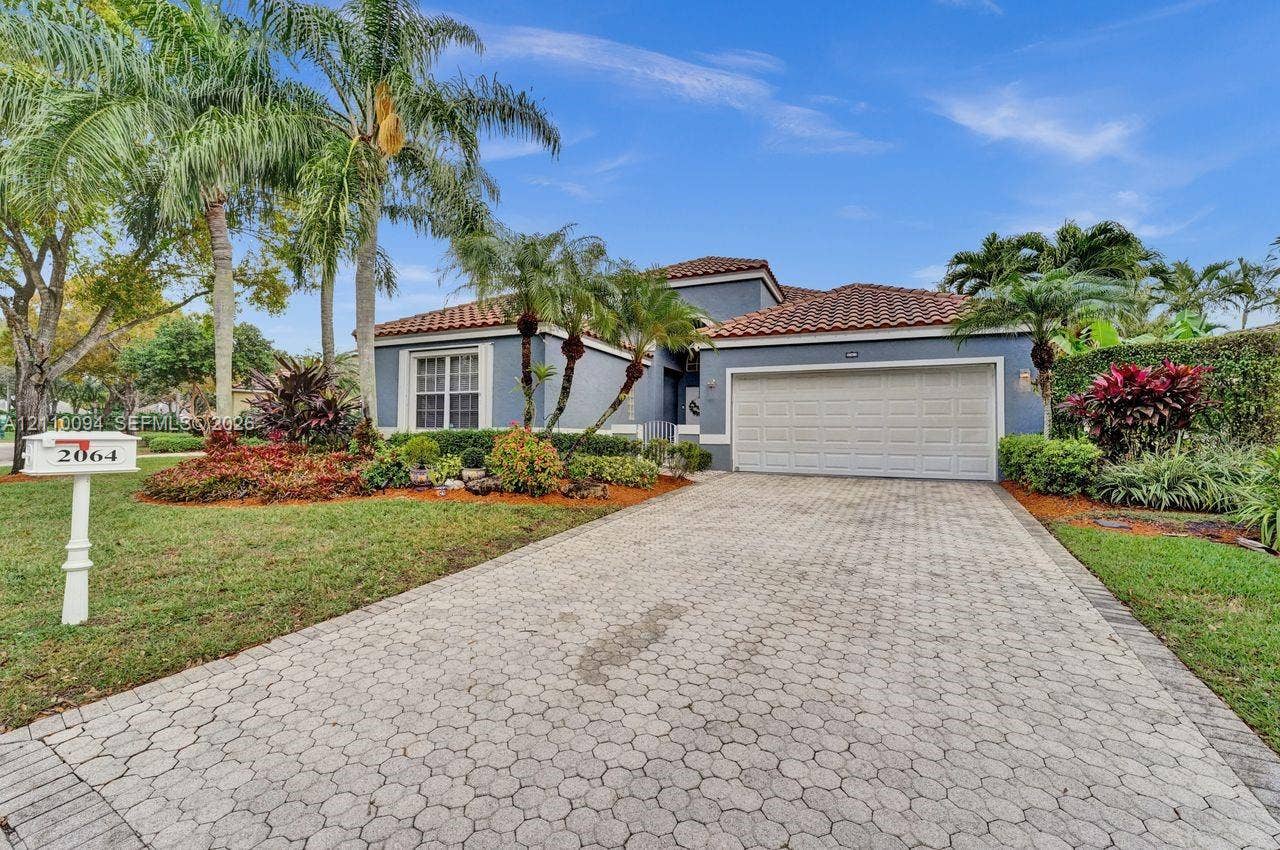 2064 Island Circle, Weston, FL 33326 Exterior facade featuring a tiled roof, multi-car garage, and paver driveway, complemented by mature tropical landscaping