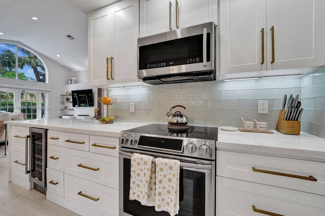 2064 Island Circle, Weston, FL 33326 Kitchen featuring white shaker-style cabinetry with brass hardware, solid surface countertops, a subway tile backsplash, stainless steel appliances, and an under-counter wine refrigerator