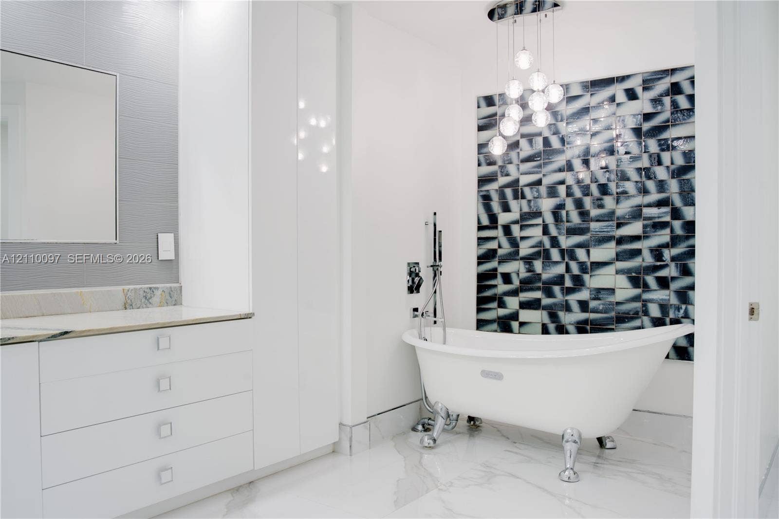 Clawfoot soaking tub with polished chrome hardware, geometric patterned accent wall, white cabinetry with integrated countertop, multi-globe pendant lighting, and large-format tile flooring