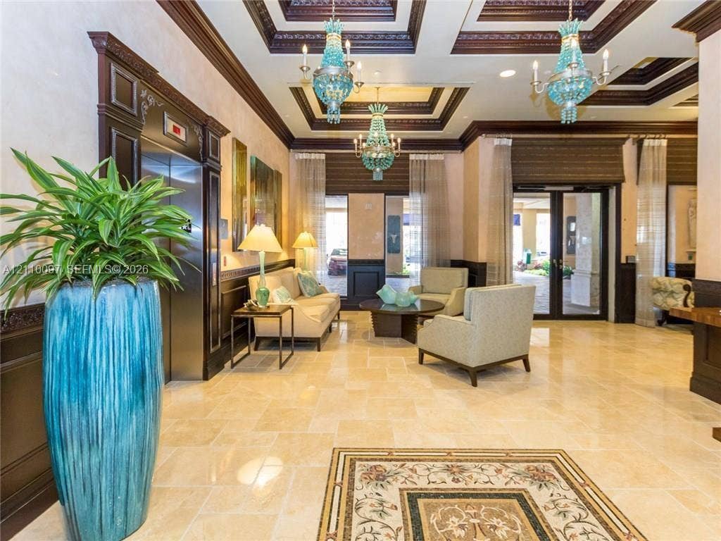 Grand entrance lobby featuring a tray ceiling with crown molding, stone tile flooring, an inlaid decorative floor medallion, ornate blue glass chandeliers, and dark wood-finished trim