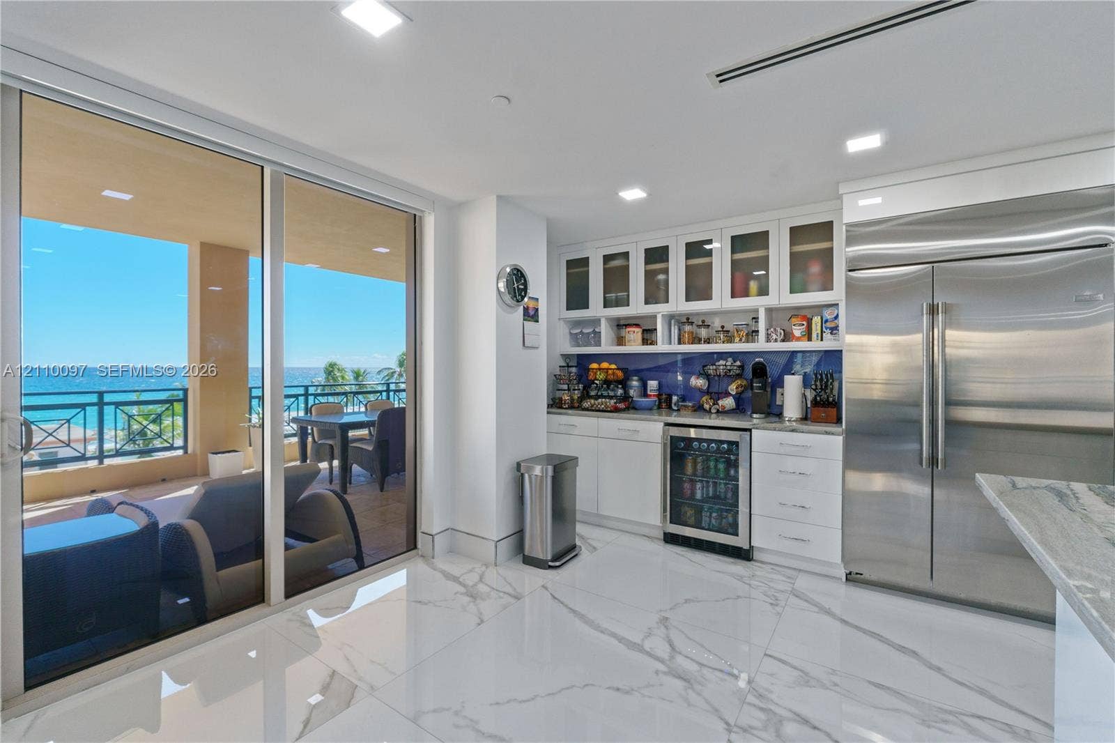 Large-format tile flooring, built-in white cabinetry with glass-front uppers, stainless steel side-by-side refrigerator, under-counter beverage cooler, and expansive floor-to-ceiling sliding glass doors opening to a covered balcony