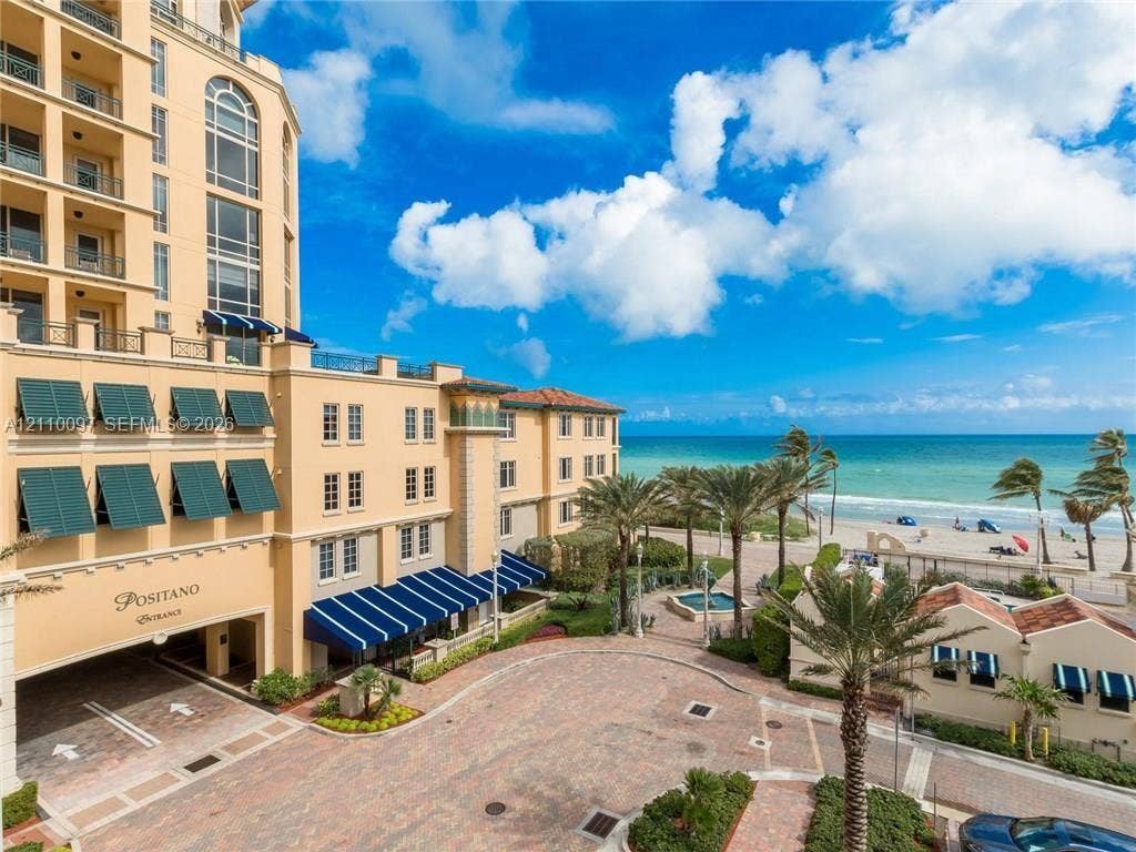 Multi-story Mediterranean-style exterior featuring a covered porte-cochre, blue fabric awnings, paver-stone driveway, and immediate proximity to the coastline