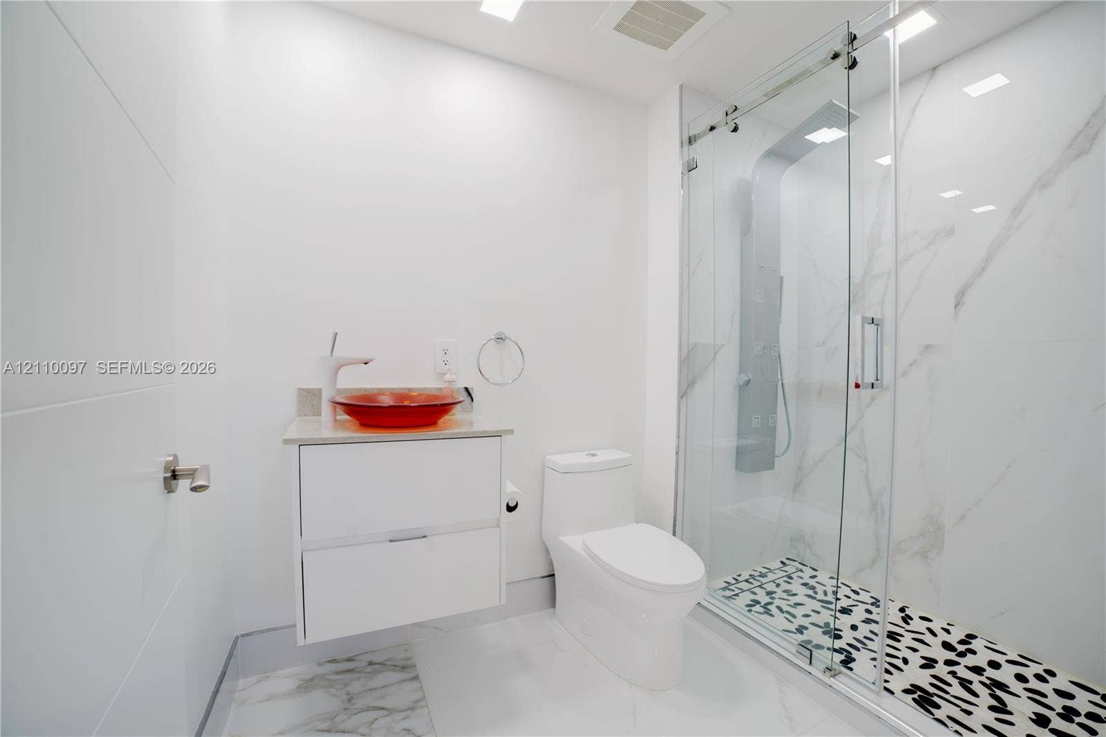 Floating white vanity with a colorful vessel sink, wall-mounted faucet, and walk-in shower featuring a glass sliding door, stone-patterned wall panels, and patterned floor tile