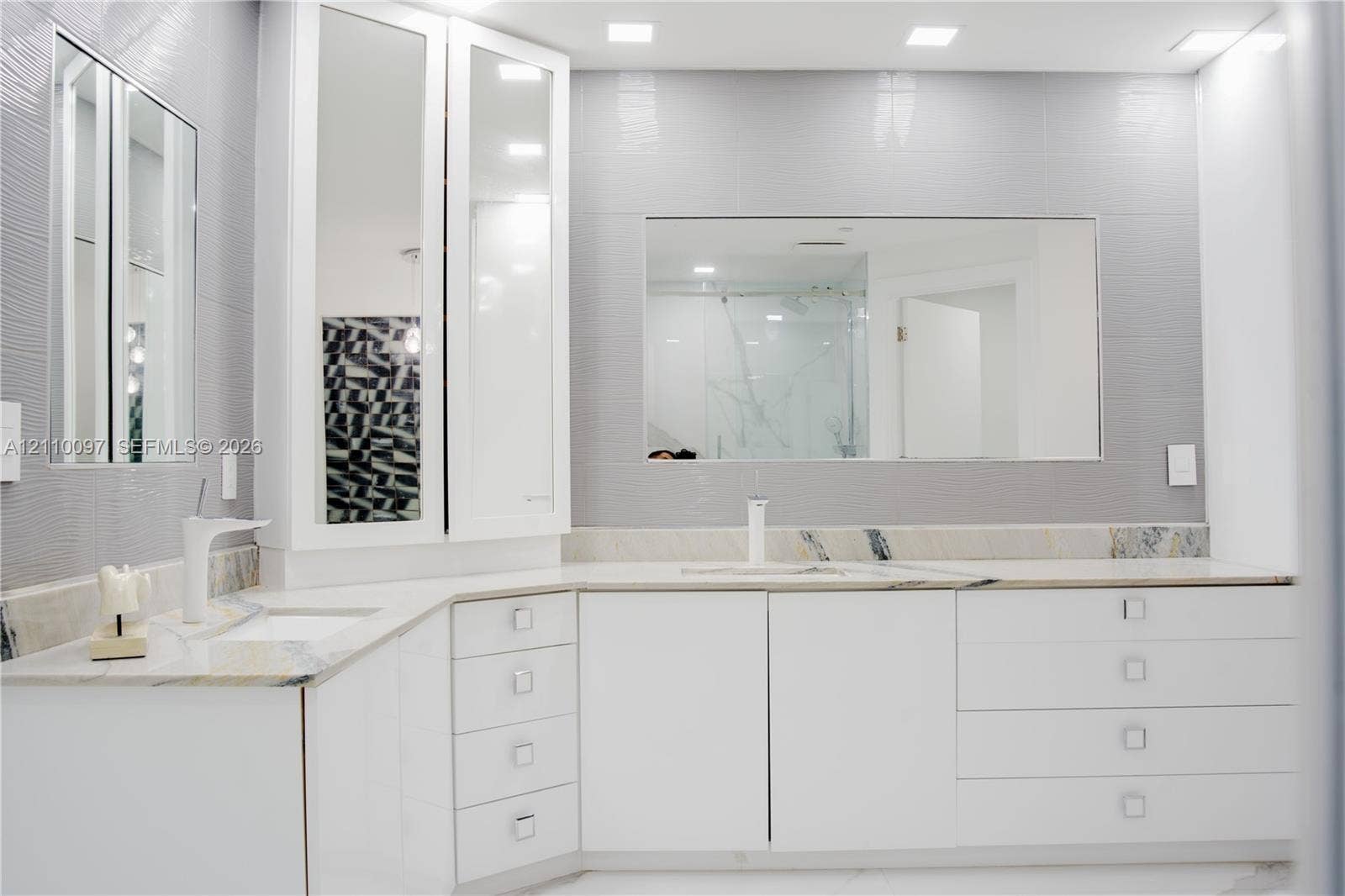 Custom white cabinetry with rectangular hardware, expansive countertop, dual recessed mirrors, and grey textured wall tiling