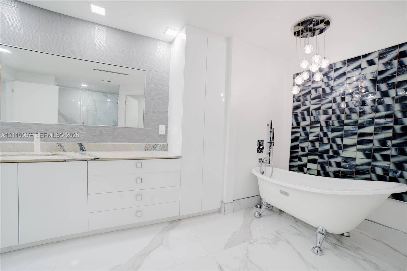 Freestanding clawfoot tub with chrome hardware, floor-to-ceiling geometric decorative wall tiling, custom white cabinetry with stone countertop, large-format veined flooring, and modern multi-globe chandelier lighting
