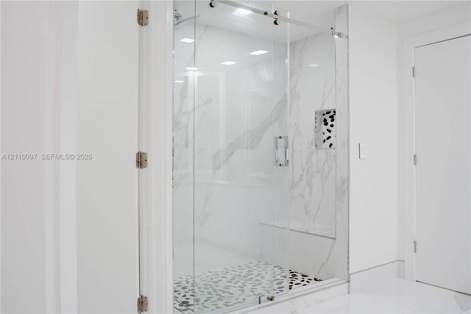 Walk-in shower featuring a frameless glass door, stone-finish wall tile with veining, pebble-style shower floor, built-in tiled bench, and an integrated recessed storage niche