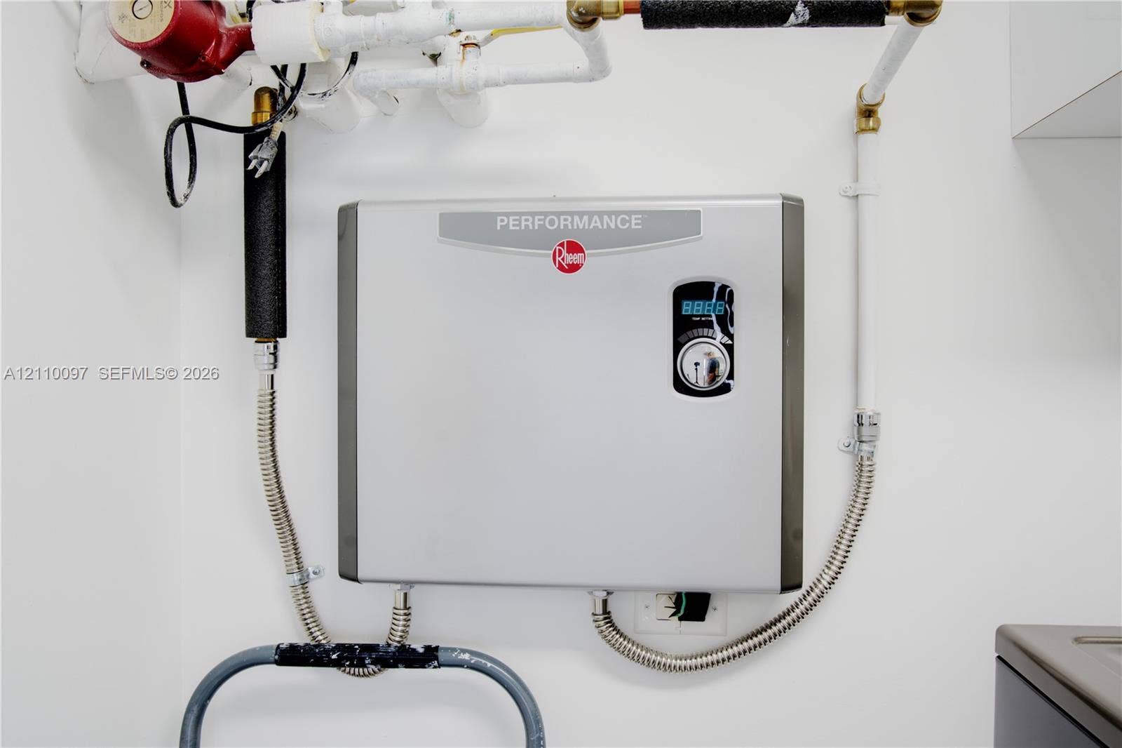 Wall-mounted tankless electric water heater featuring a digital temperature display and metallic finish casing with professional-grade plumbing connections