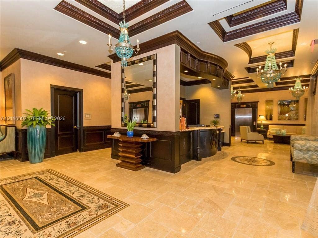 Grand lobby featuring coffered ceilings with dark wood molding, natural stone flooring with inlaid medallion accents, custom built-in reception desk, decorative crystal chandeliers, and wall-mounted mirrors
