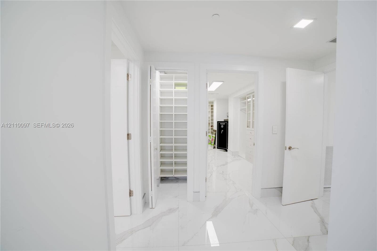 Large-format stone-finish flooring with white and grey veining throughout, featuring multiple walk-in storage areas with custom built-in shelving, white slab-style doors, and recessed lighting