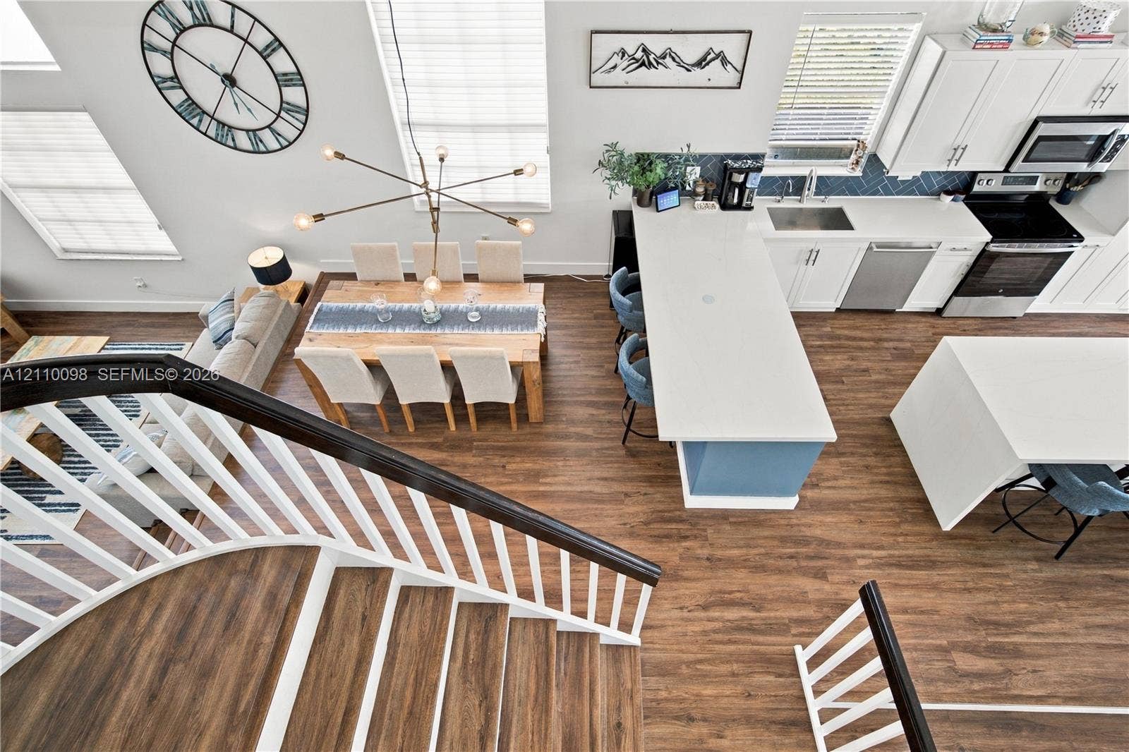 Open floor plan featuring wood-finish flooring, a kitchen with a quartz-topped island, white cabinetry, herringbone backsplash, and a staircase with a dark-stained handrail