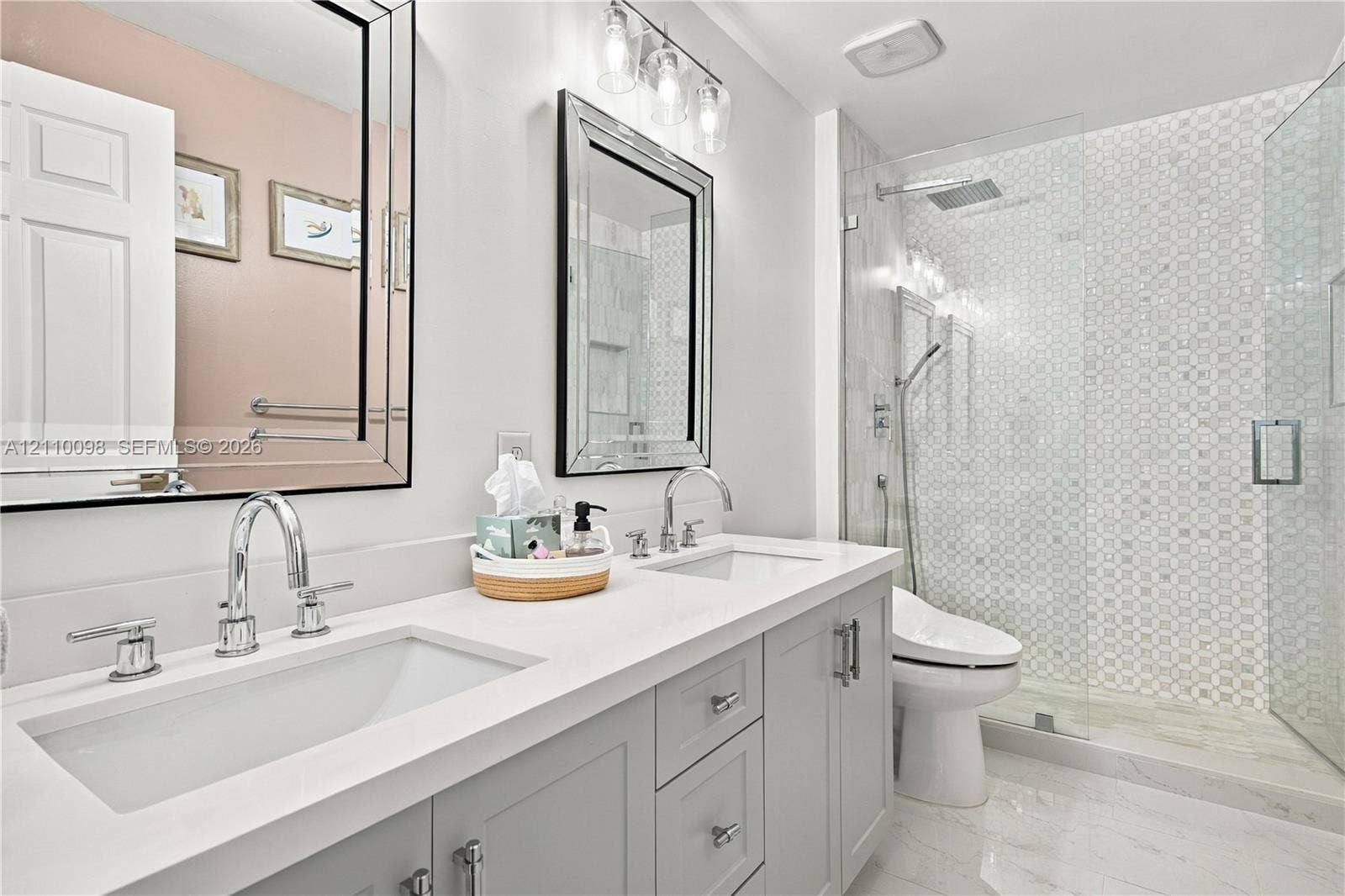 Dual-vanity cabinetry with a light-toned solid surface countertop, undermount sinks, polished chrome hardware, walk-in glass shower with decorative wall tile, and framed wall mirrors