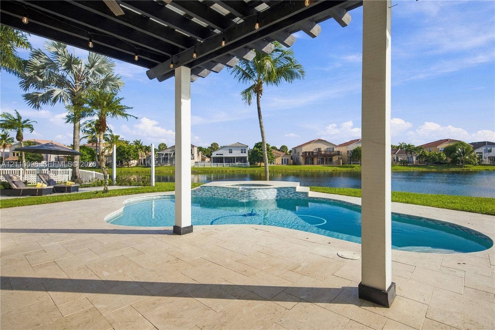 Covered patio featuring a wood-beam pergola, stone-paver decking, an in-ground pool with a raised spa, and waterfront grounds