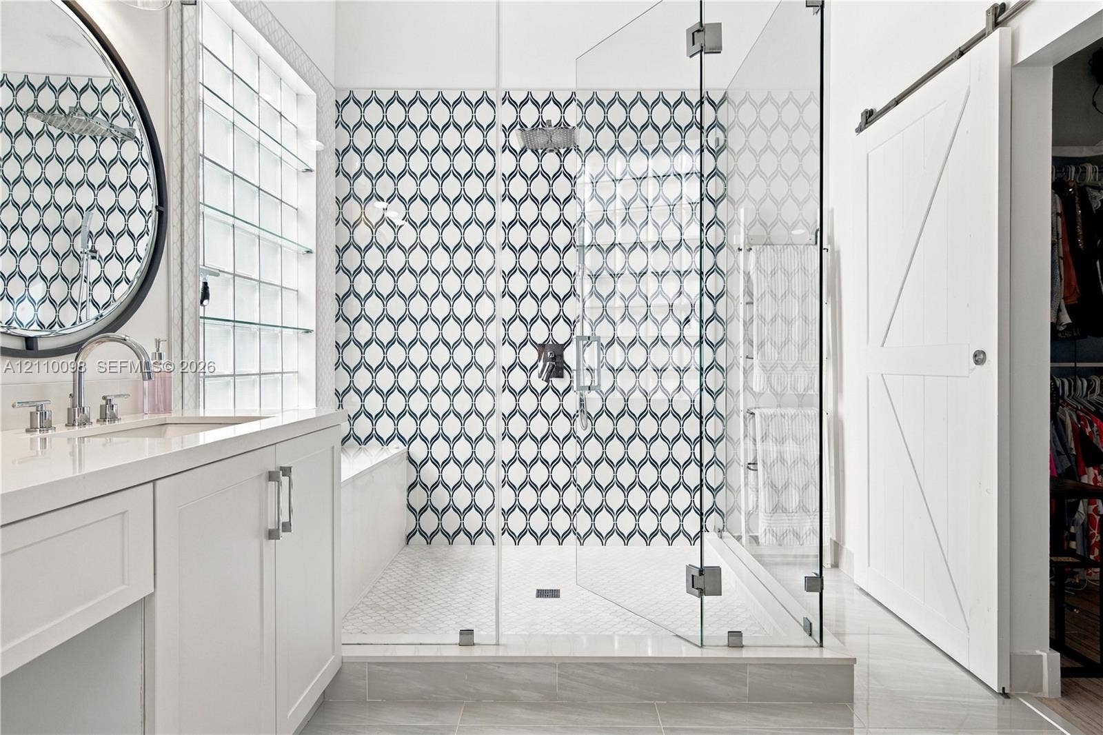Walk-in shower featuring glass paneling and patterned tile surround, white cabinetry with light-toned countertop, round wall-mounted mirror, and white sliding barn door