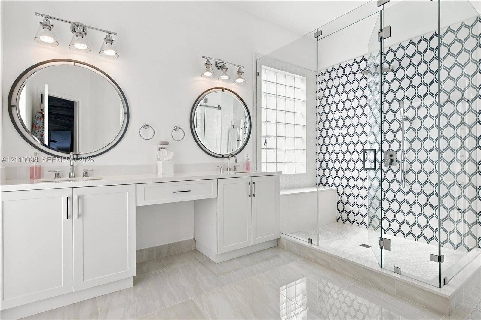 Double vanity featuring white cabinetry, oval mirrors, dual three-light fixtures, and a walk-in glass-enclosed shower with patterned tile surround