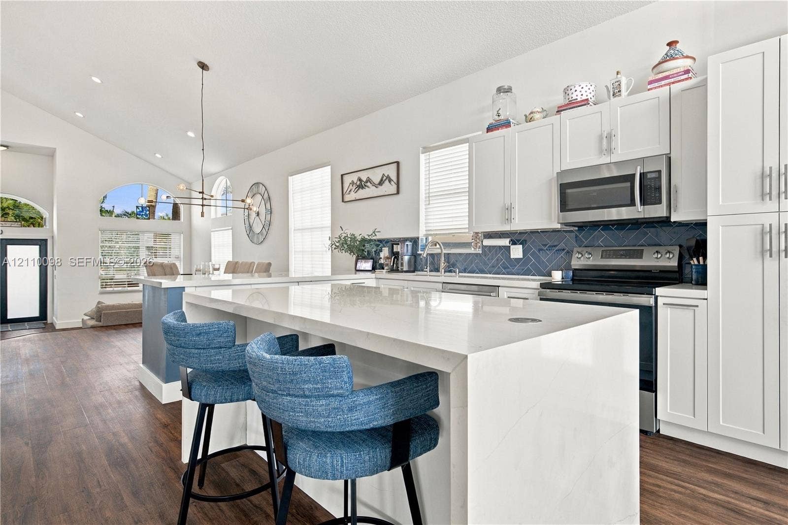 Kitchen featuring a quartz-topped island with waterfall edge, white shaker-style cabinetry, blue herringbone-pattern tile backsplash, stainless steel appliances, and wood-finish flooring