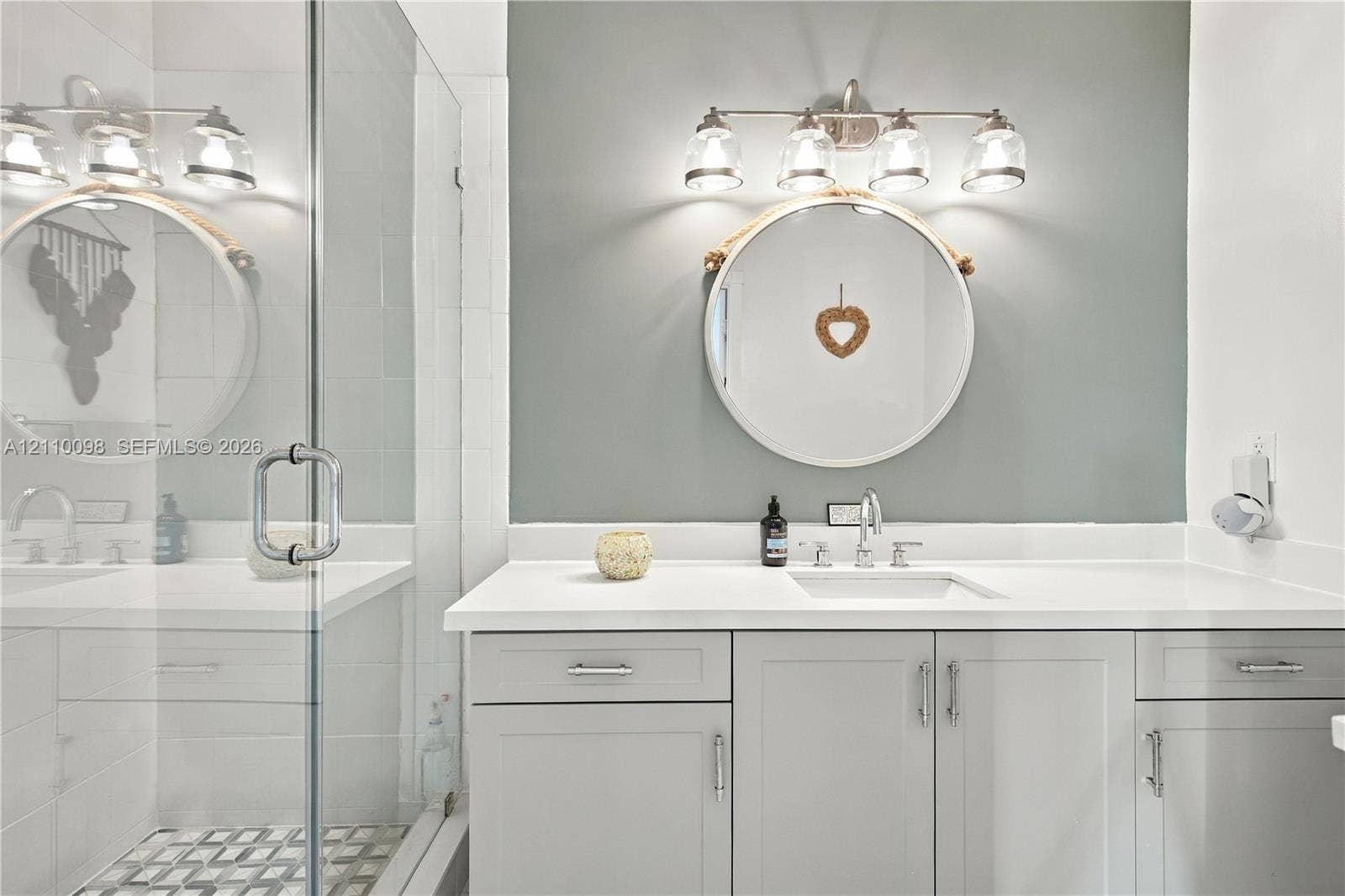 Bathroom featuring a vanity with light gray cabinetry, white solid-surface countertop, rectangular undermount sink, four-light vanity fixture, and a glass-enclosed shower with patterned tile flooring