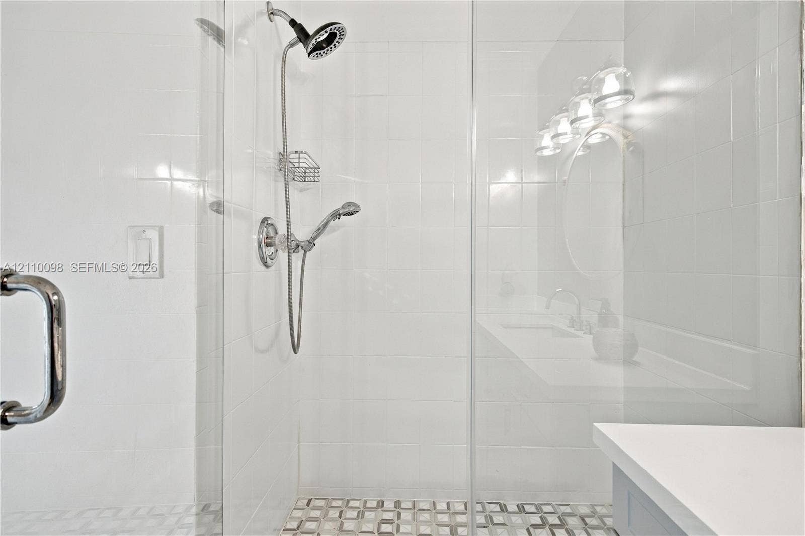 Walk-in glass shower enclosure featuring white tile walls, a handheld showerhead, built-in wire shelving, and a patterned tile floor
