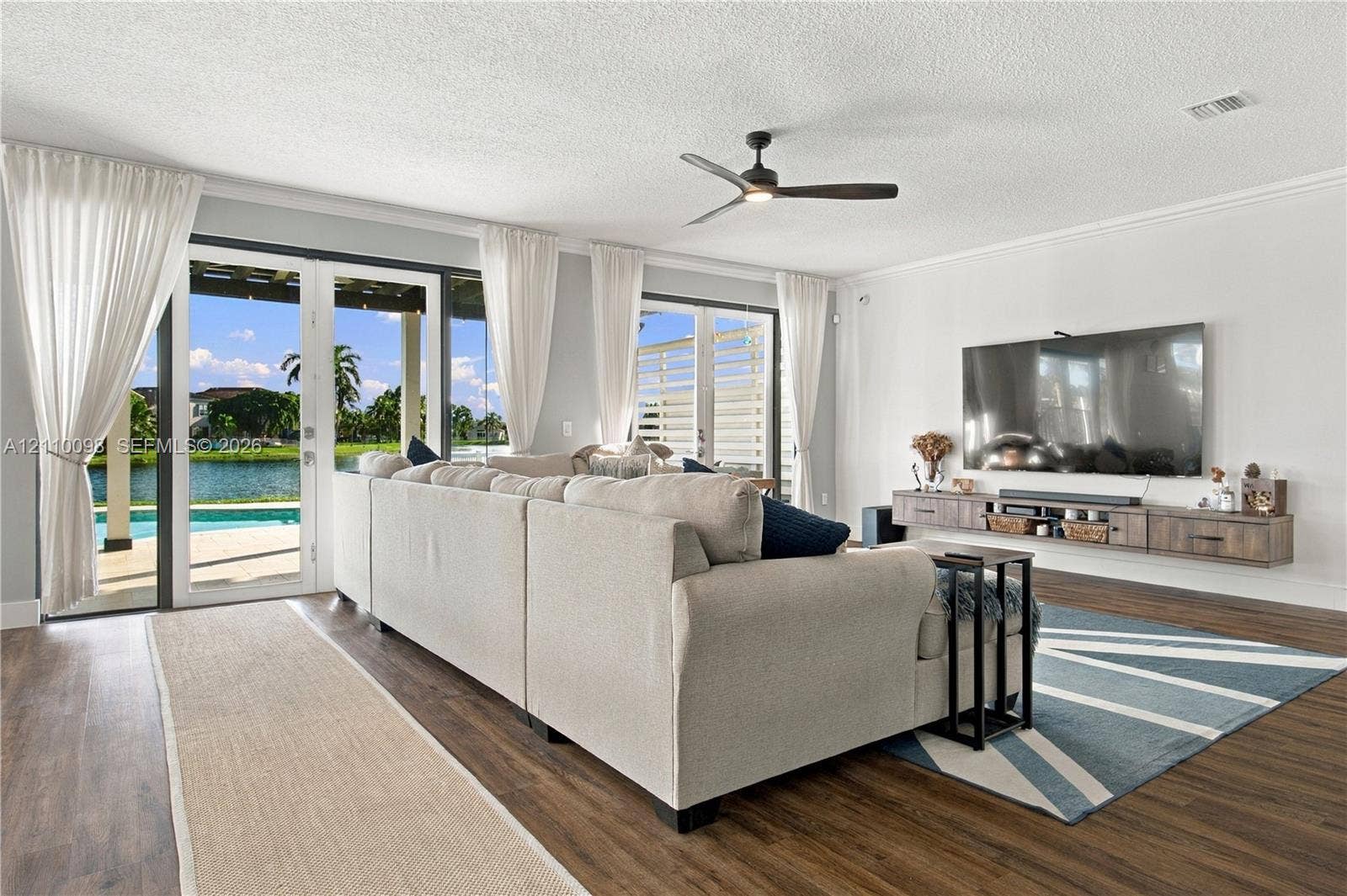 Living area featuring wood-finish flooring, a wall-mounted floating media console, a ceiling fan, and double glass sliding doors opening to a water-front patio