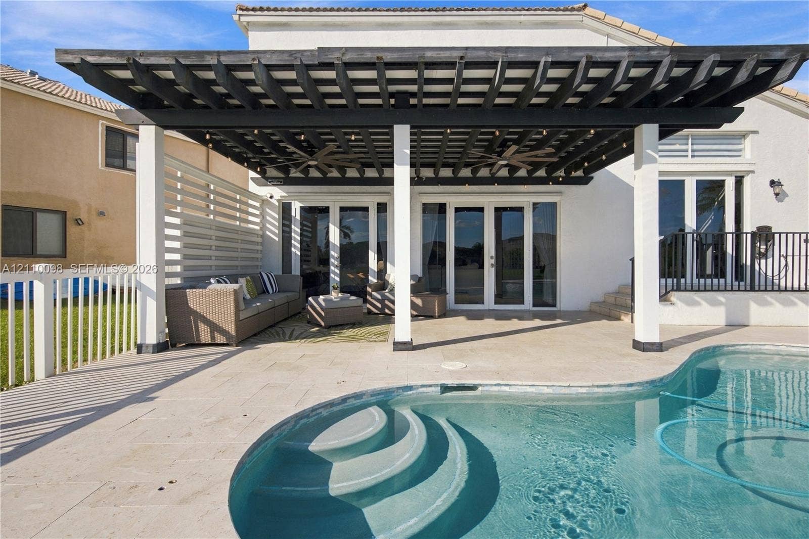 Covered patio featuring a dark-finish pergola, integrated ceiling fans, stone-finish decking, and a curved pool with recessed stairs