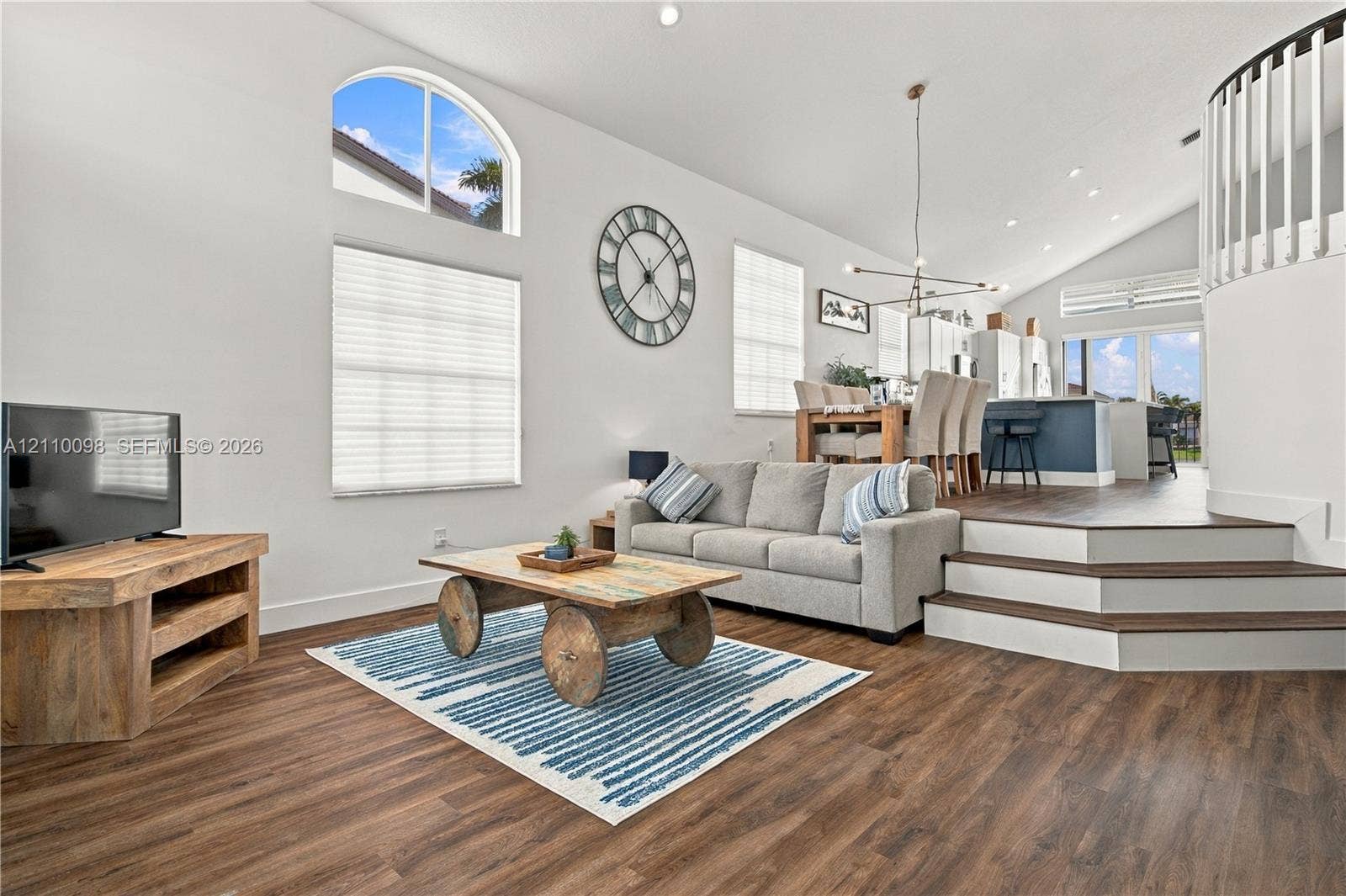 Open-concept living space featuring vaulted ceilings, wood-finish flooring, an arched window, and a multi-level staircase with white balusters