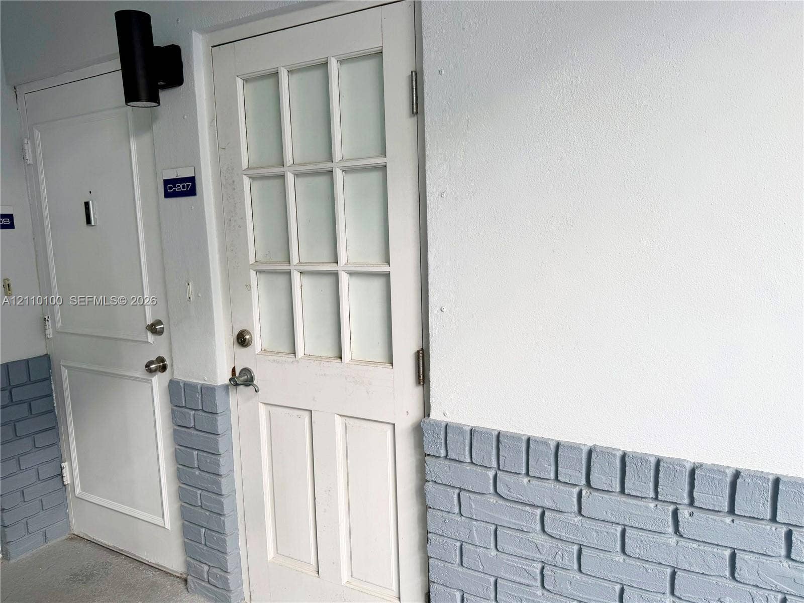 Exterior entry featuring a multi-light glass pane door, paneled solid door, cylindrical wall-mounted sconce, and painted brick wainscoting