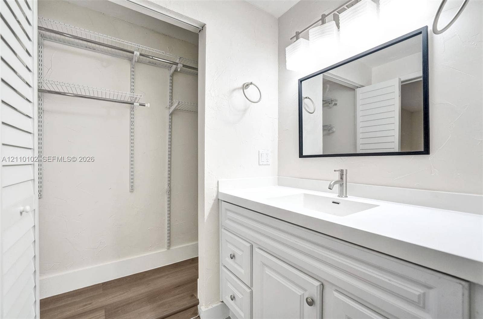 1224 NE 13th Avenue, #2, Fort Lauderdale, Fort Lauderdale, FL 33304 Bathroom vanity featuring a white cabinet, integrated sink, solid-surface countertop, and rectangular mirror with a black frame, accompanied by an open closet with wire shelving and wood-finish flooring