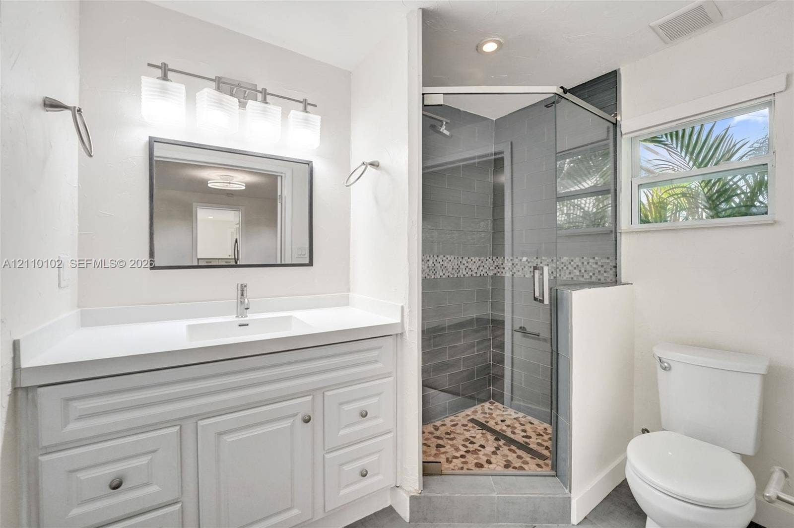 1224 NE 13th Avenue, #2, Fort Lauderdale, Fort Lauderdale, FL 33304 White cabinetry vanity with a clean-lined countertop, rectangular undermount sink, four-light vanity fixture, corner glass-enclosed shower featuring decorative tile accents and pebble-style flooring, and a single-hung window