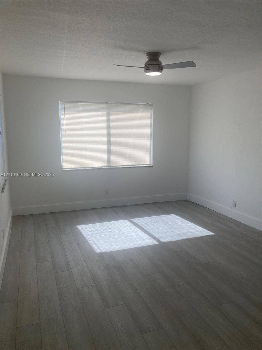 Neutral-toned interior room featuring wood-finish flooring, a ceiling-mounted light fixture with a fan, and a single window with fitted horizontal blinds