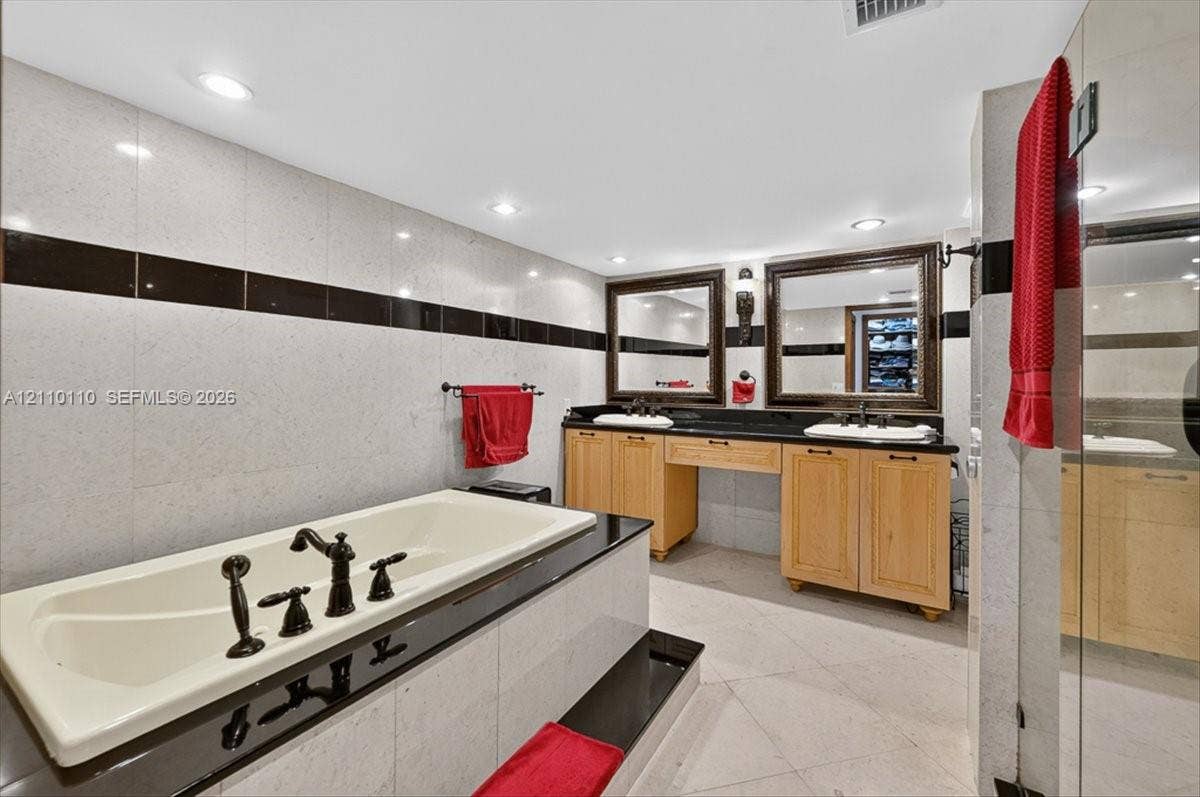 2751 S Ocean Drive, #801N, Hollywood, FL 33019 Dual-vanity cabinetry with light wood finish, integrated stone-surface countertop, recessed lighting, drop-in soaking tub with dark-finish hardware, and tile wall accents