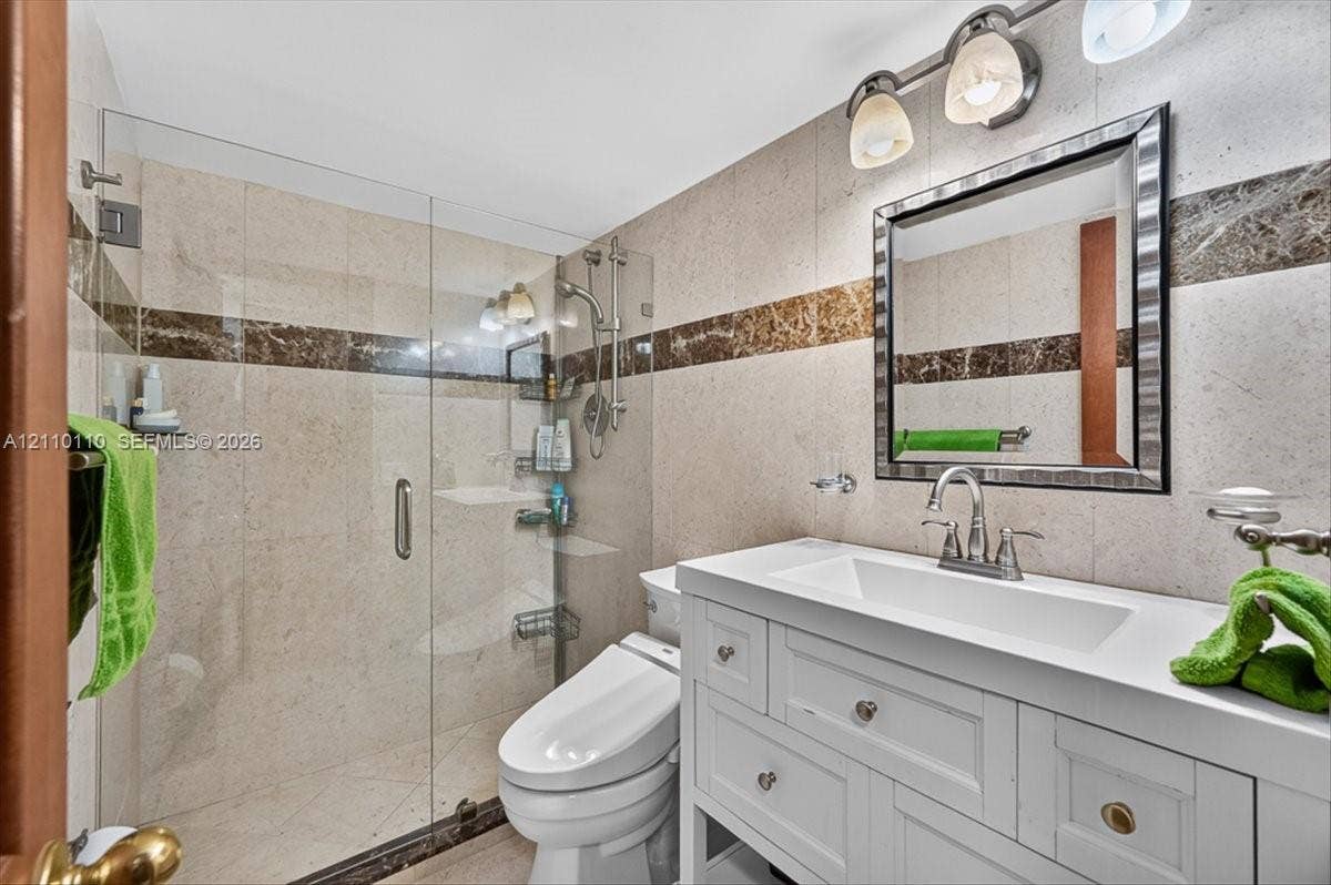 2751 S Ocean Drive, #801N, Hollywood, FL 33019 White vanity with integrated rectangular basin and metal hardware, walk-in glass-enclosed shower, stone wall tiling with decorative inlay, and wall-mounted vanity lighting
