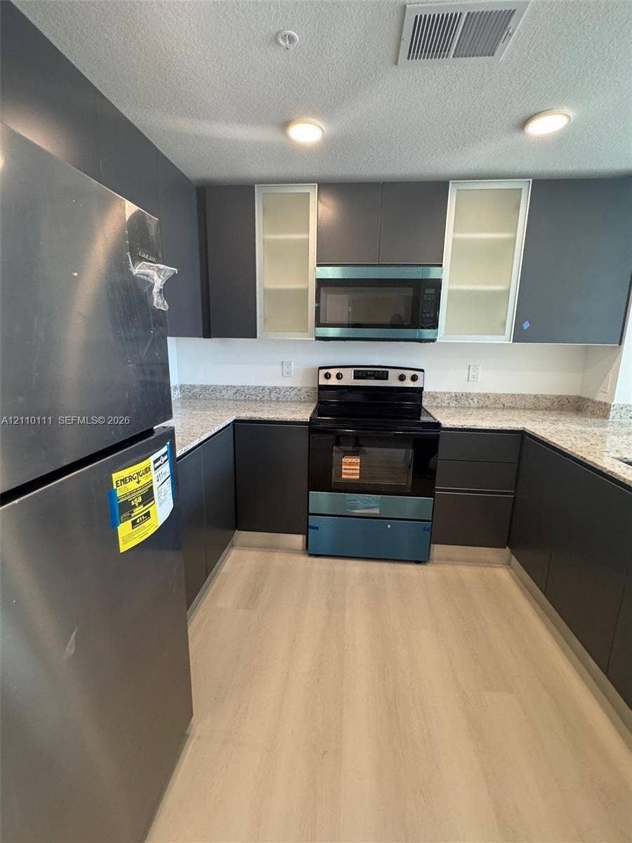 5563 Millenia Park Boulevard, #315, Orlando, Orlando, FL 32839 Kitchen featuring flat-panel dark cabinetry, glass-fronted upper cabinets, stone-composite countertops, stainless steel appliances, and wood-finish flooring