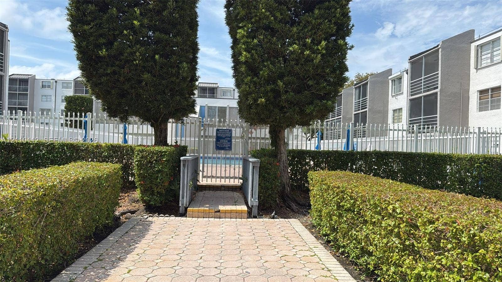 14185 SW 87th Street, #A110, Miami, FL 33183 Common area swimming pool enclosed by metal fencing, featuring a paver stone walkway and mature hedgerow landscaping