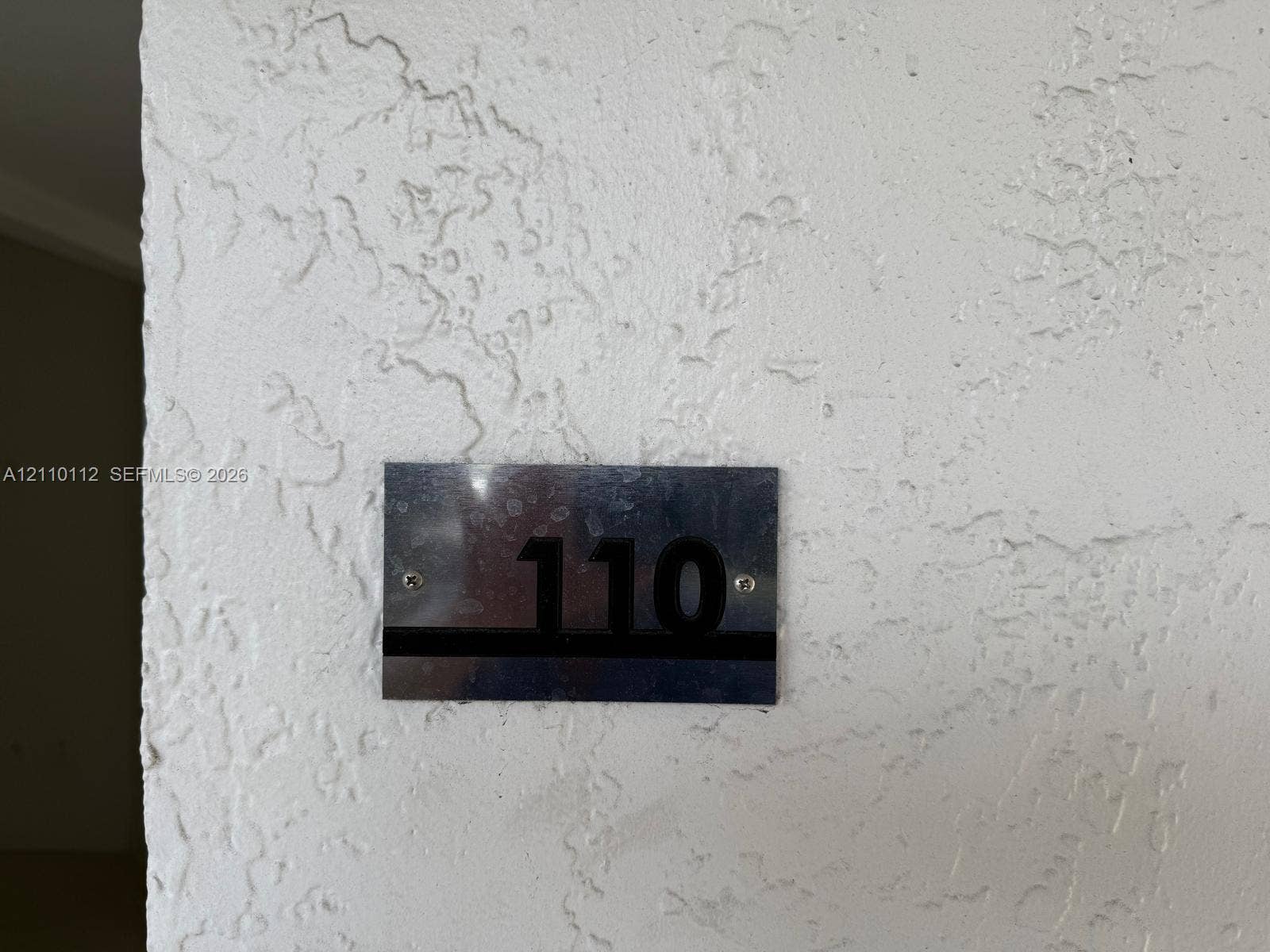 14185 SW 87th Street, #A110, Miami, FL 33183 Metallic, rectangular unit identification plaque with raised black numbering mounted on a textured plaster wall surface