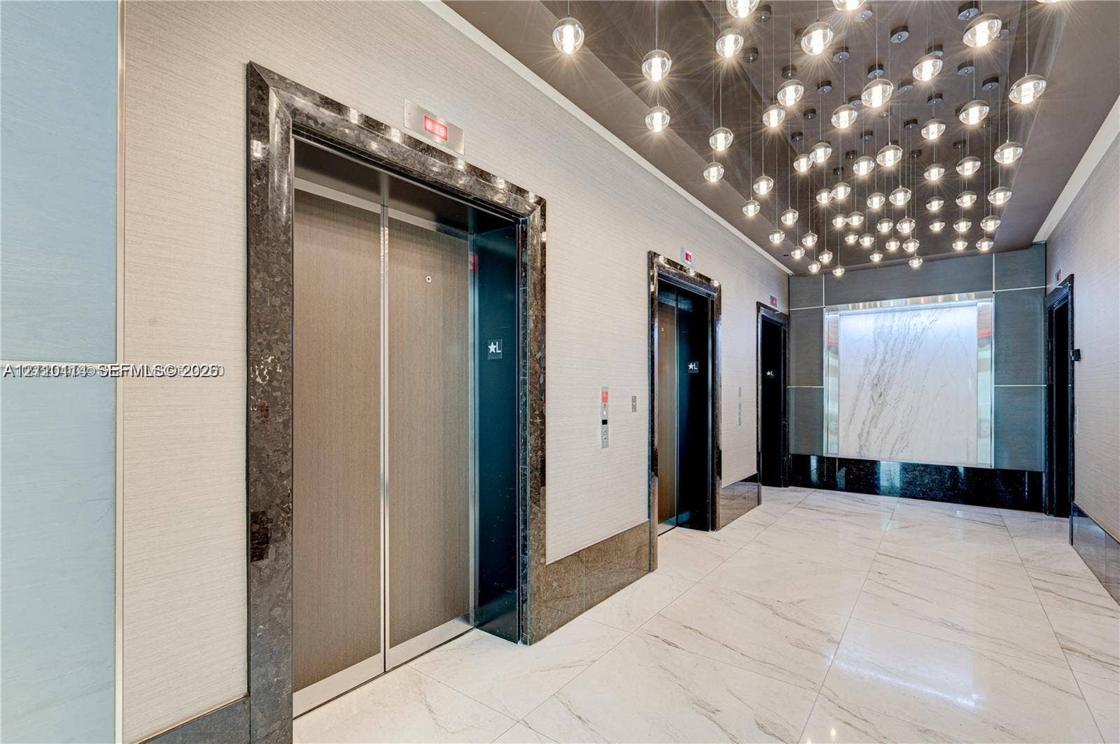 950 Brickell Bay Drive, #5108, Miami, Miami, FL 33131 Elevator lobby featuring metallic elevator doors with stone-trim surrounds, large-format tile flooring, a decorative cluster of globe pendant lighting, and a backlit stone accent wall