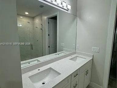 3304 11 Street W, #0, Lehigh Acres, FL 33971 Dual-sink vanity with a solid-surface countertop, white shaker-style cabinetry, a full-width mirror, and a glass-enclosed shower with tiled surround