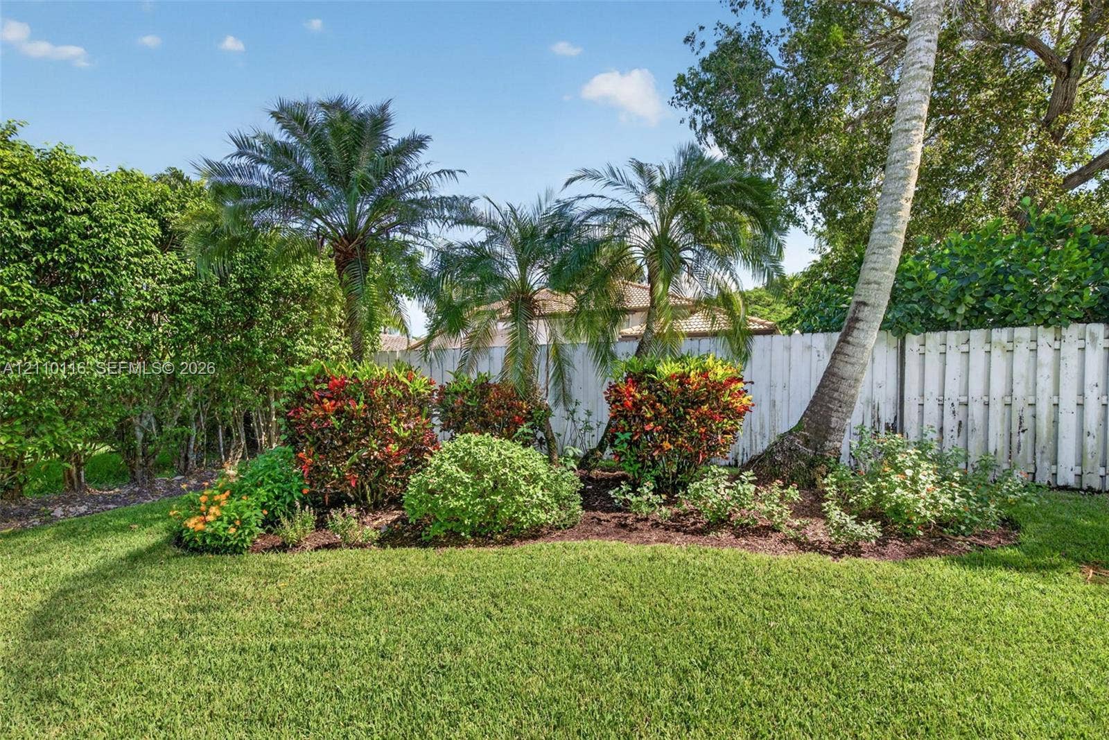 Backyard featuring a manicured lawn, established palm trees, ornamental shrubbery, and wood-panel fencing