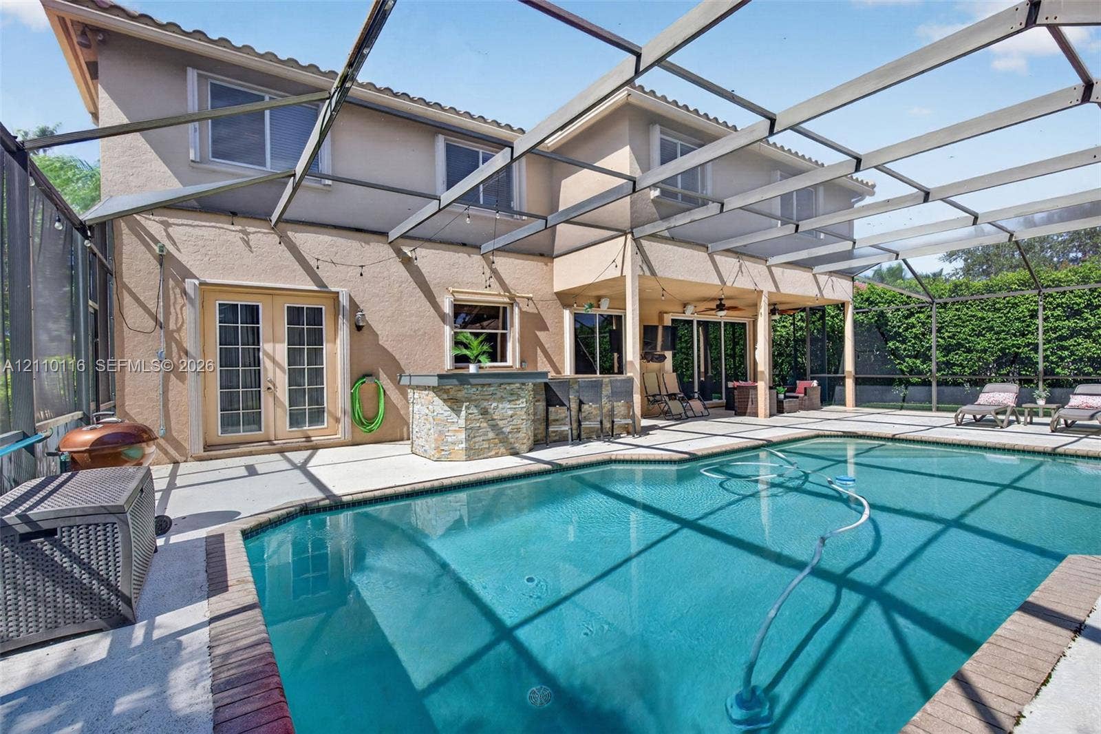 In-ground swimming pool enclosed by a screen lanai, featuring an exterior stone-veneer bar, covered patio space, and two-story stucco exterior