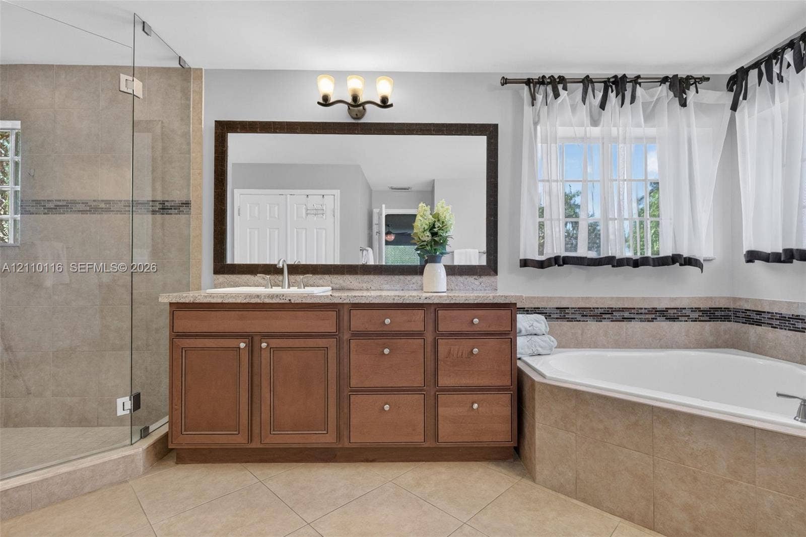 Bathroom featuring wood-finish cabinetry, stone countertop, corner soaking tub with tile surround, glass-enclosed shower, and large framed wall mirror