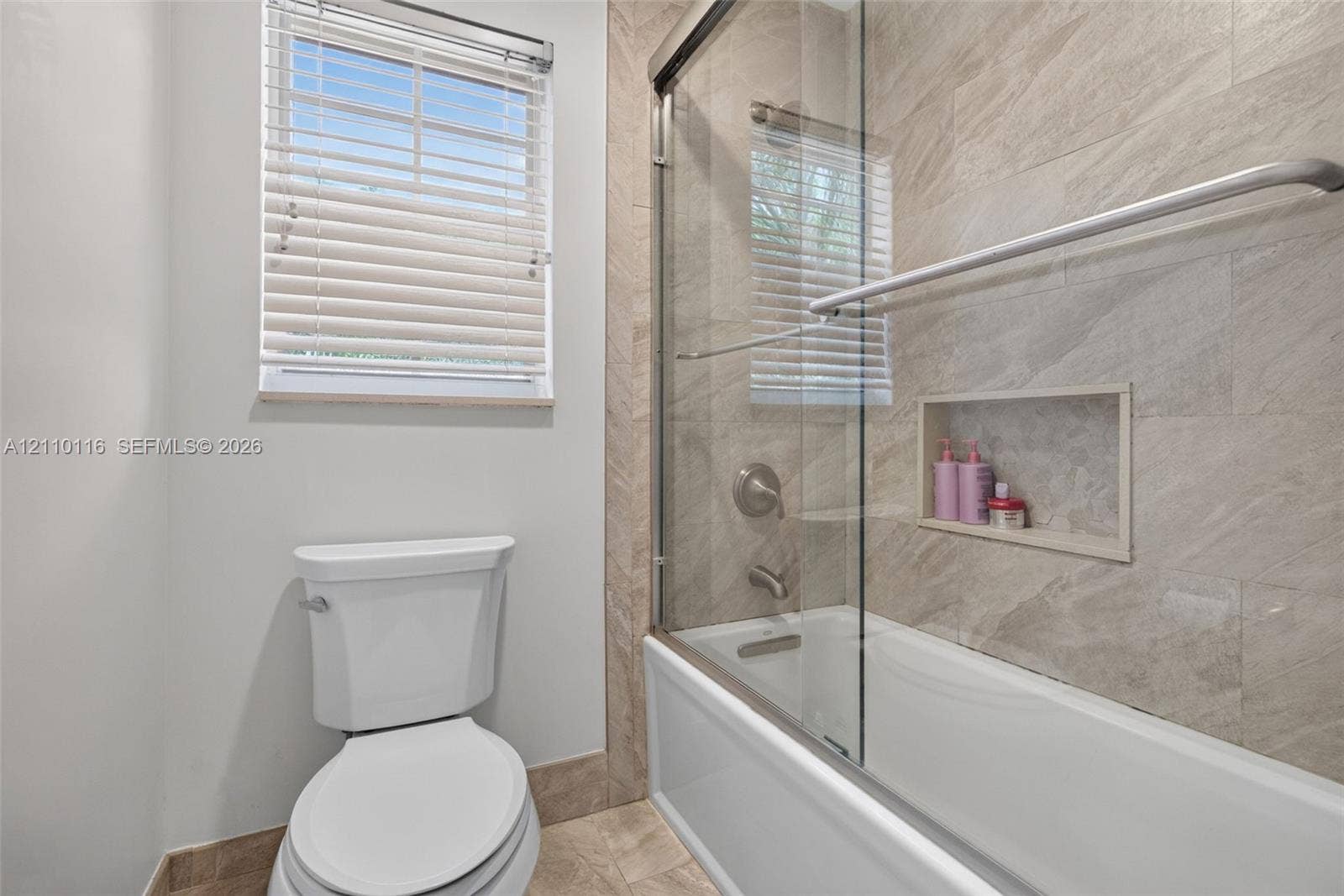 Bathroom featuring a sliding glass shower door, recessed wall niche, neutral stone-tile surround, and window with blinds