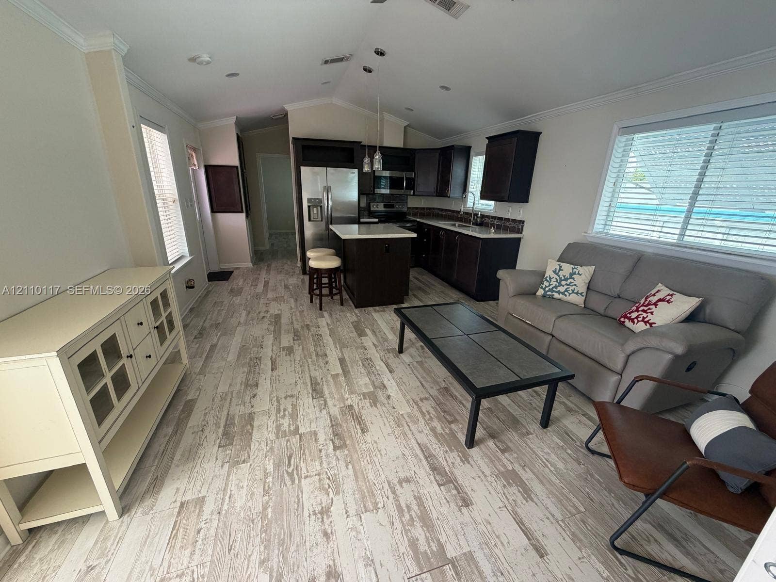 Open-concept area featuring vaulted ceilings, wood-finish flooring, dark-toned cabinetry, a central island with countertop, and stainless steel kitchen appliances