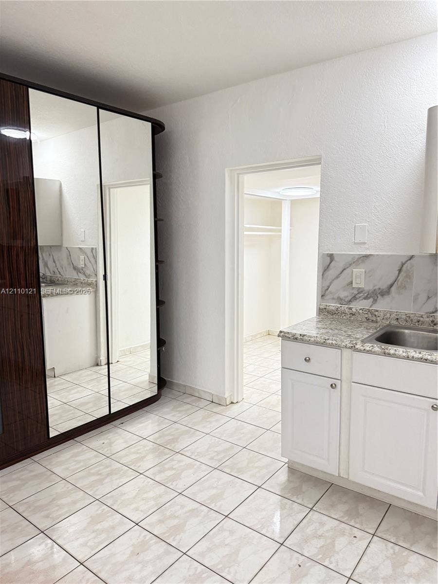Built-in floor-to-ceiling mirrored wardrobe, white cabinetry with stone-finish countertops, stainless steel undermount sink, stone-patterned backsplash, and ceramic tile flooring